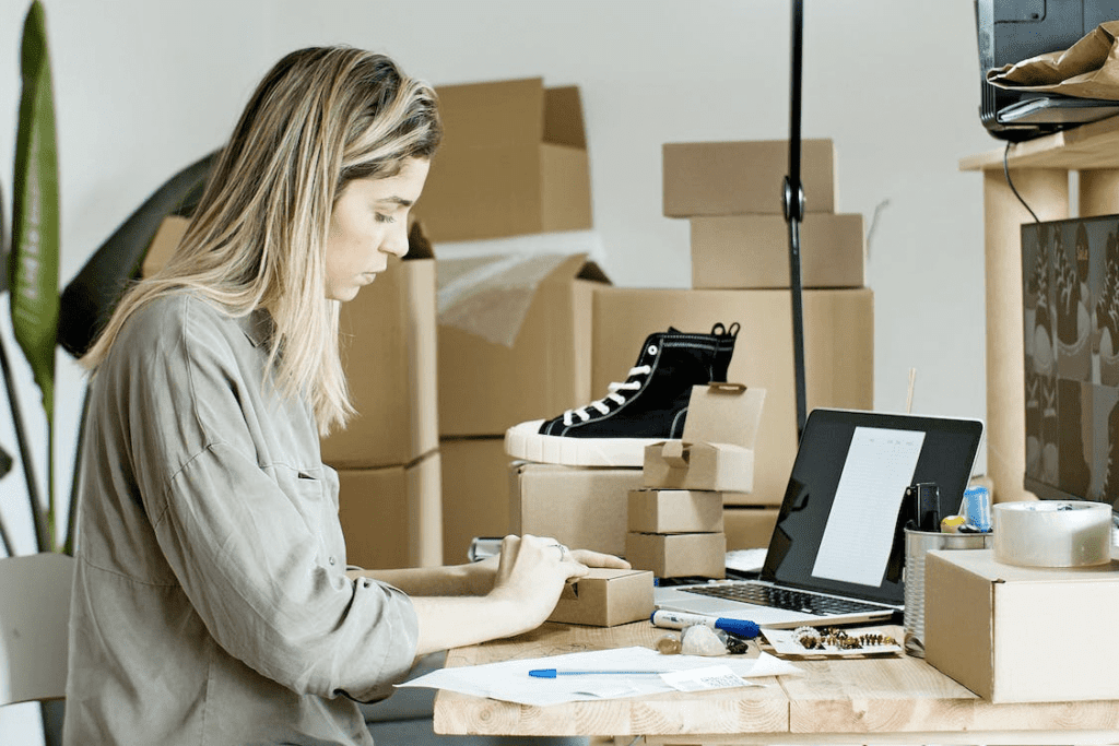 A woman working at a desk with boxes and using a laptop for WooCommerce.